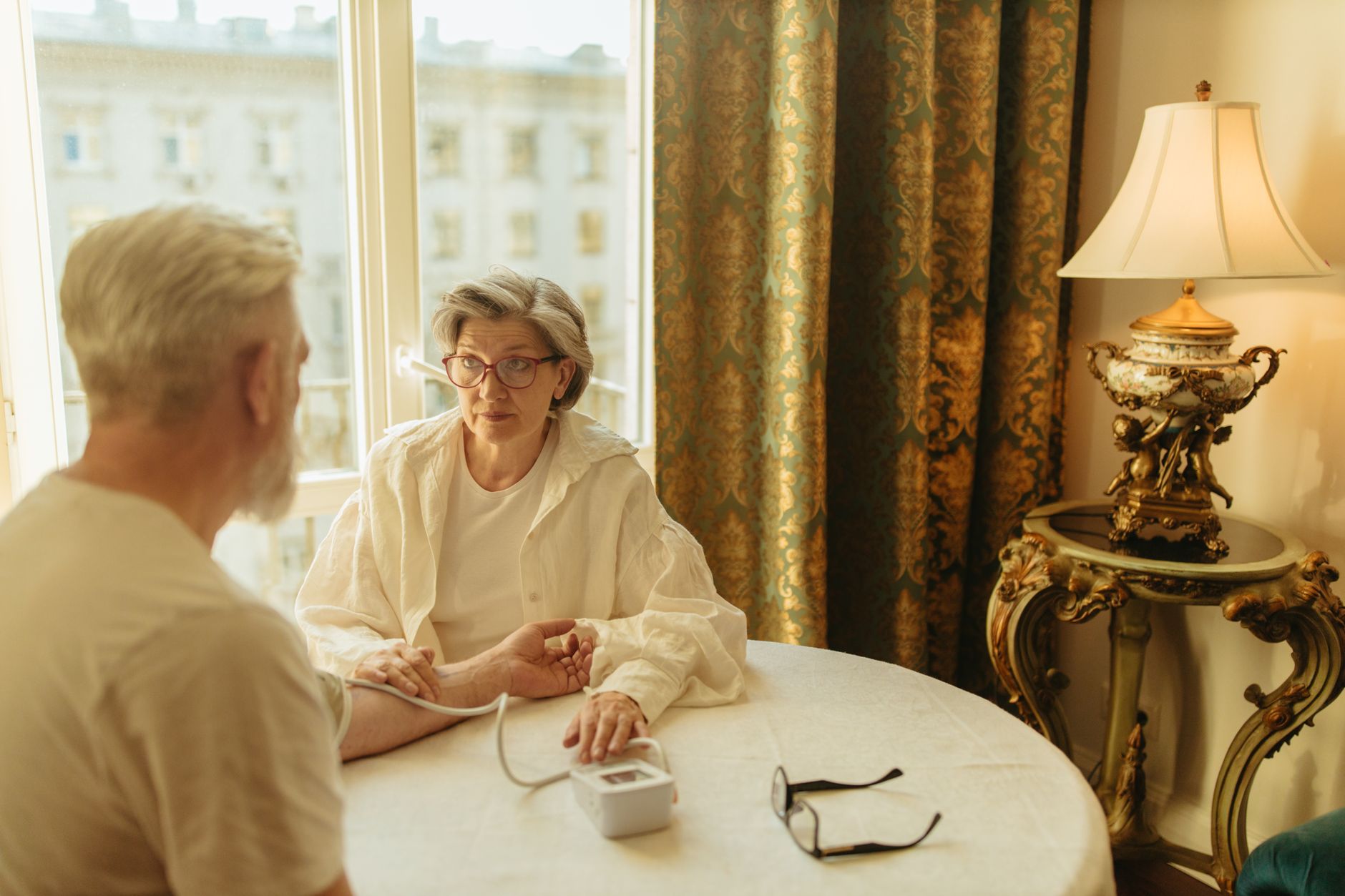An older woman checks an older man's blood pressure at a table by a sunlit window.