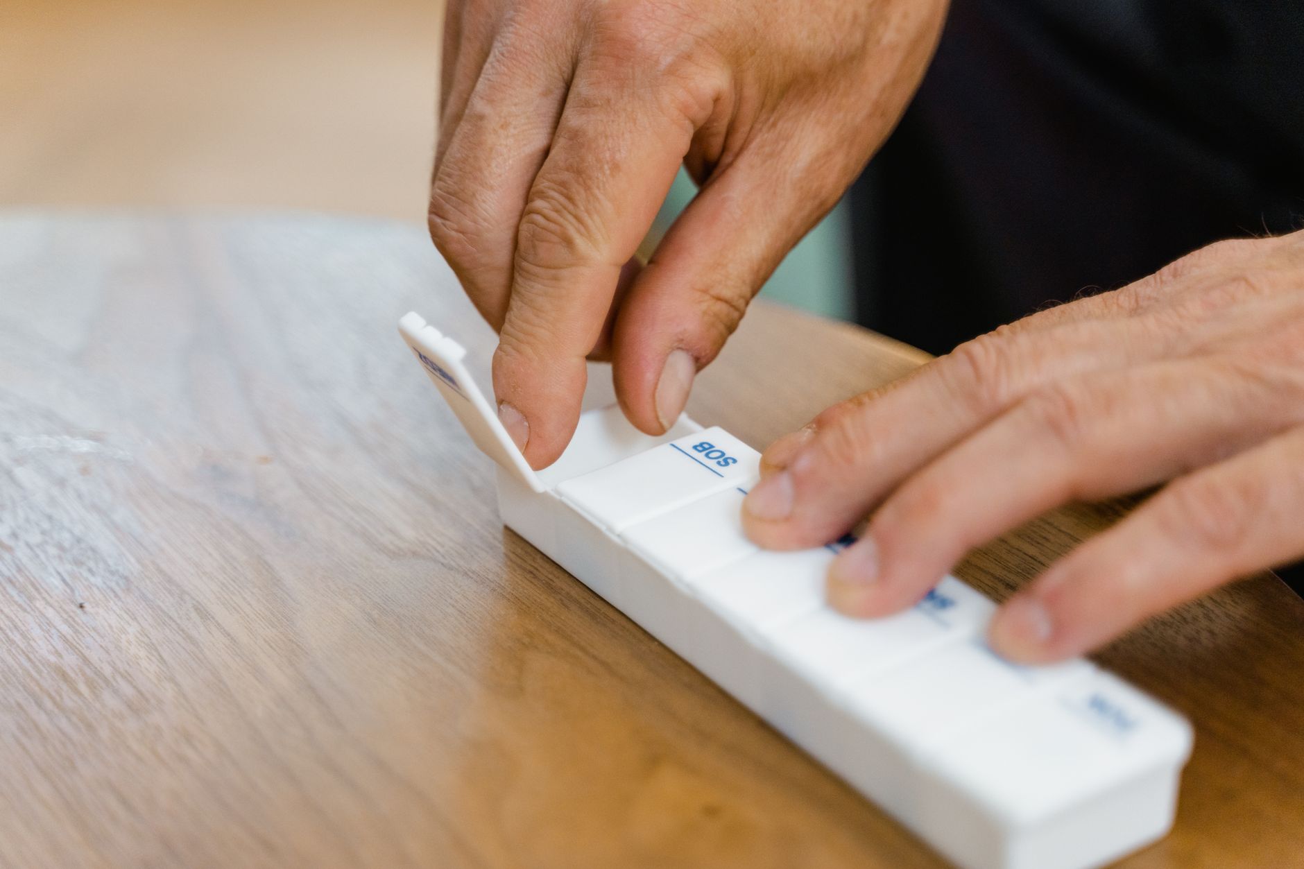 A pair of older hands sorts tablets into a weekly pill organizer on a wooden table.