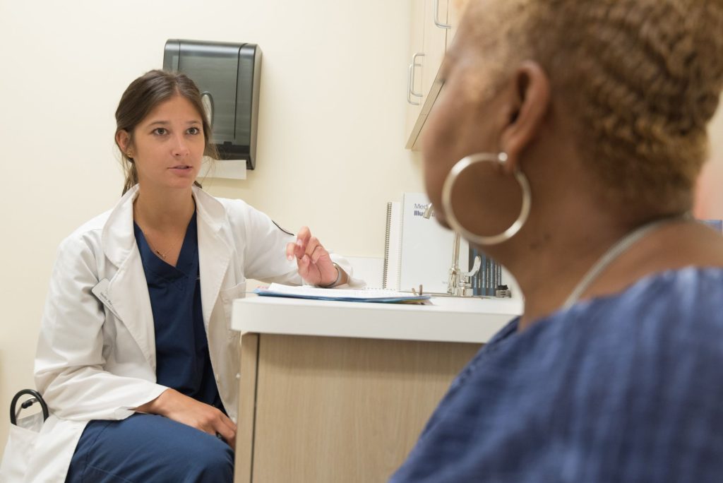 A clinician in a white coat talks with an older patient in a bright doctor's office exam room.