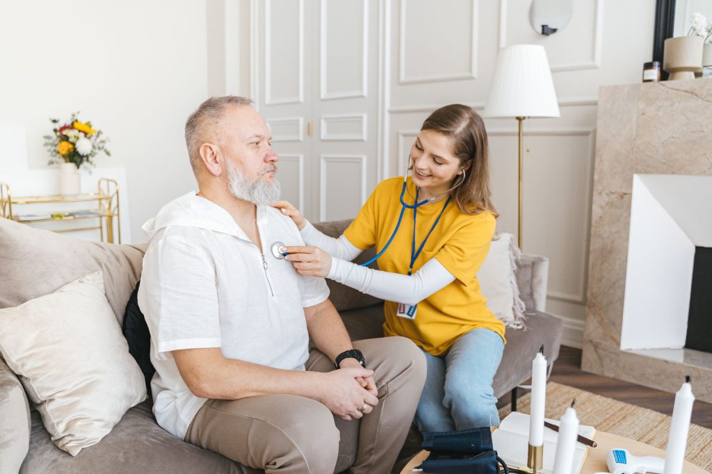A clinician listens to an older man's chest with a stethoscope during a home visit in his living room.