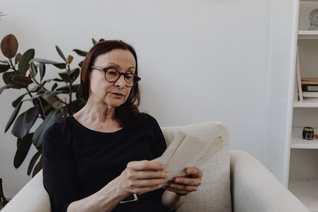 An older woman sits in an armchair at home, reading a letter through her glasses.