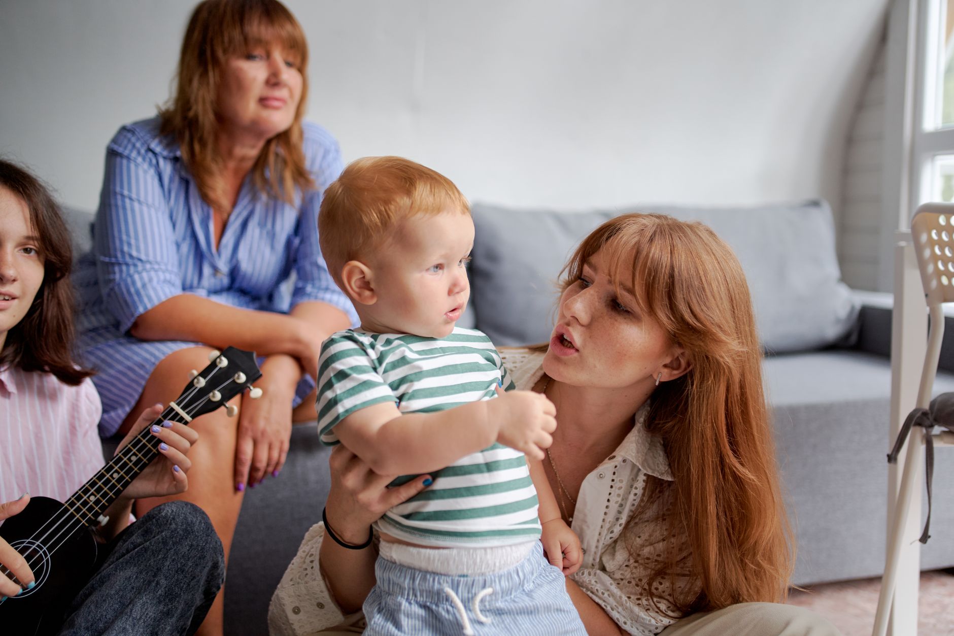 A grandmother, a young mother holding her toddler, and a teenager with a ukulele spend time together by the sofa at home.