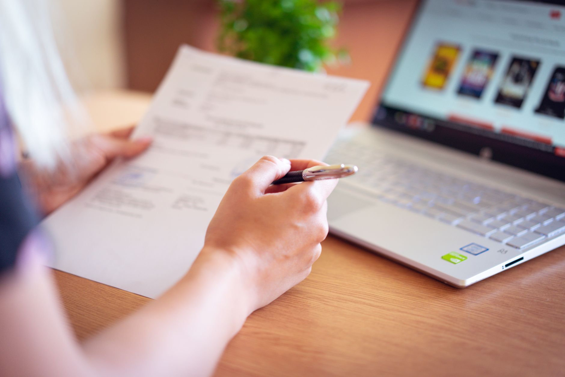Close-up of a person's hands holding a printed document and a pen beside an open laptop.
