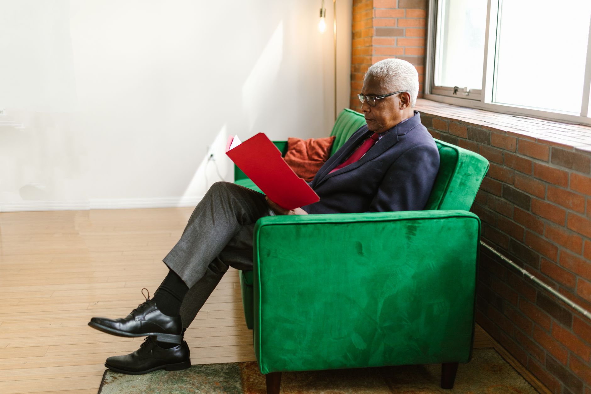 An older man sits in an armchair beside a window, reading a document in a folder.