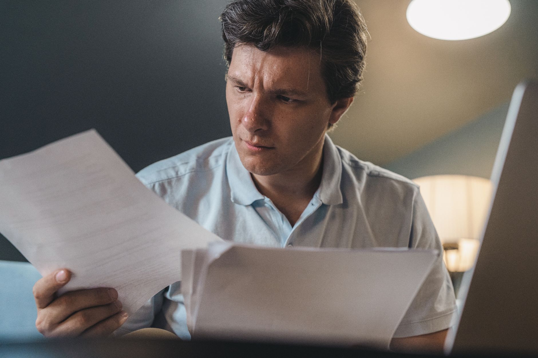 A man sits reading through printed documents at his desk, a laptop open beside him.
