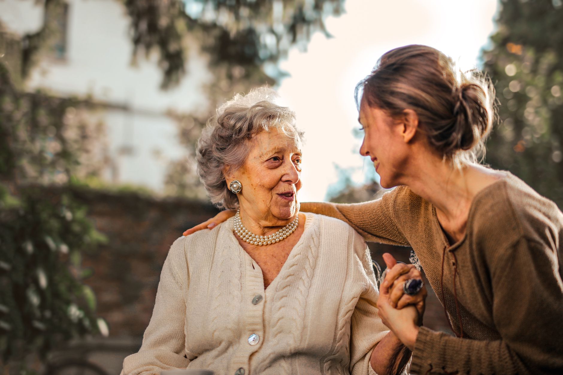 An adult daughter and her elderly mother sit talking together outdoors in a garden.