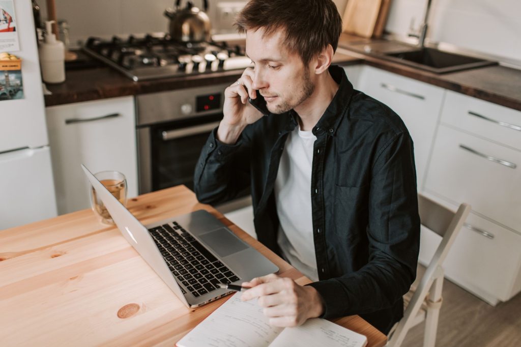 A young man talks on the phone at his kitchen table while making notes beside his laptop.