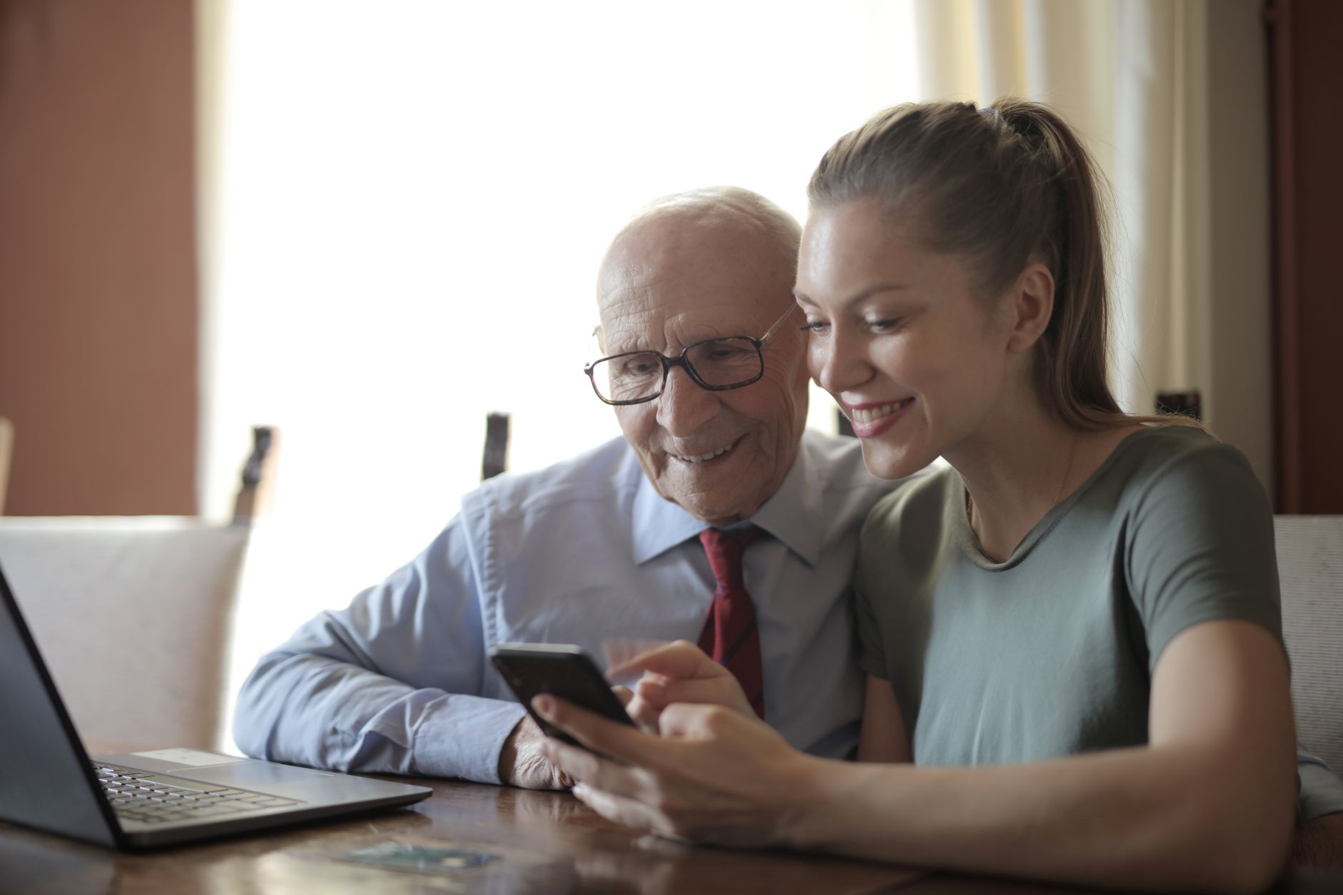 A younger woman and an older man in glasses look at a phone together at a table, a laptop open beside them.