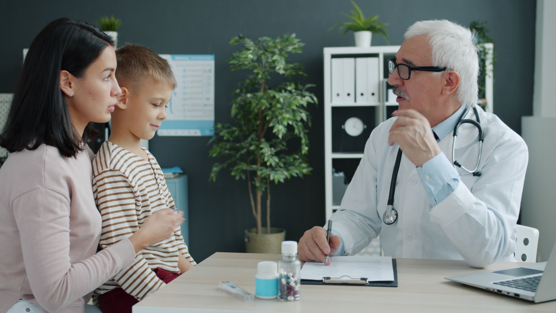 A doctor talks with a mother and her young son across a desk in a modern clinic office.