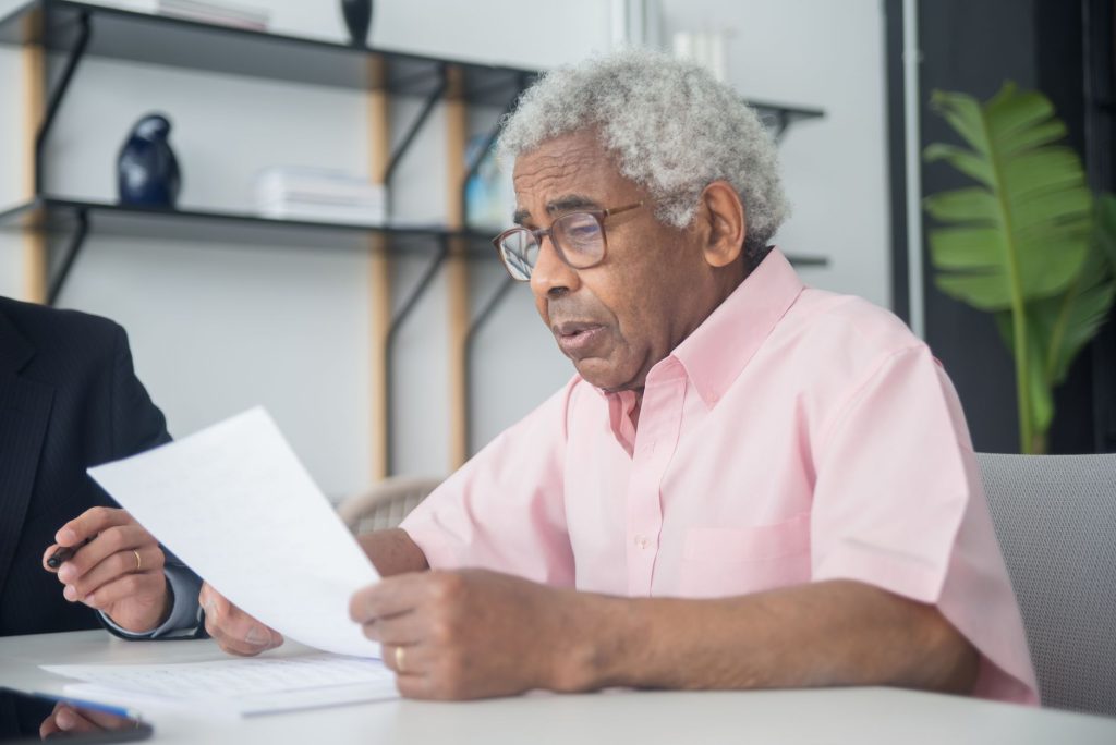 An older man in glasses reads a document closely at a table, with another person seated beside him.