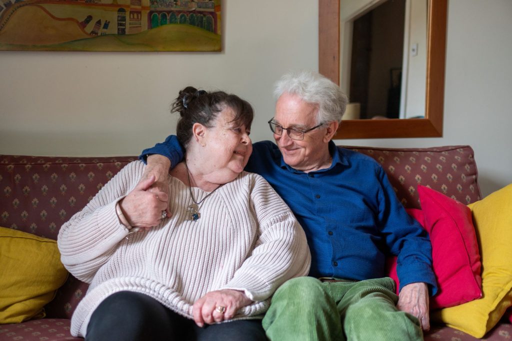 An older couple sit close together on a sofa at home, smiling at each other.
