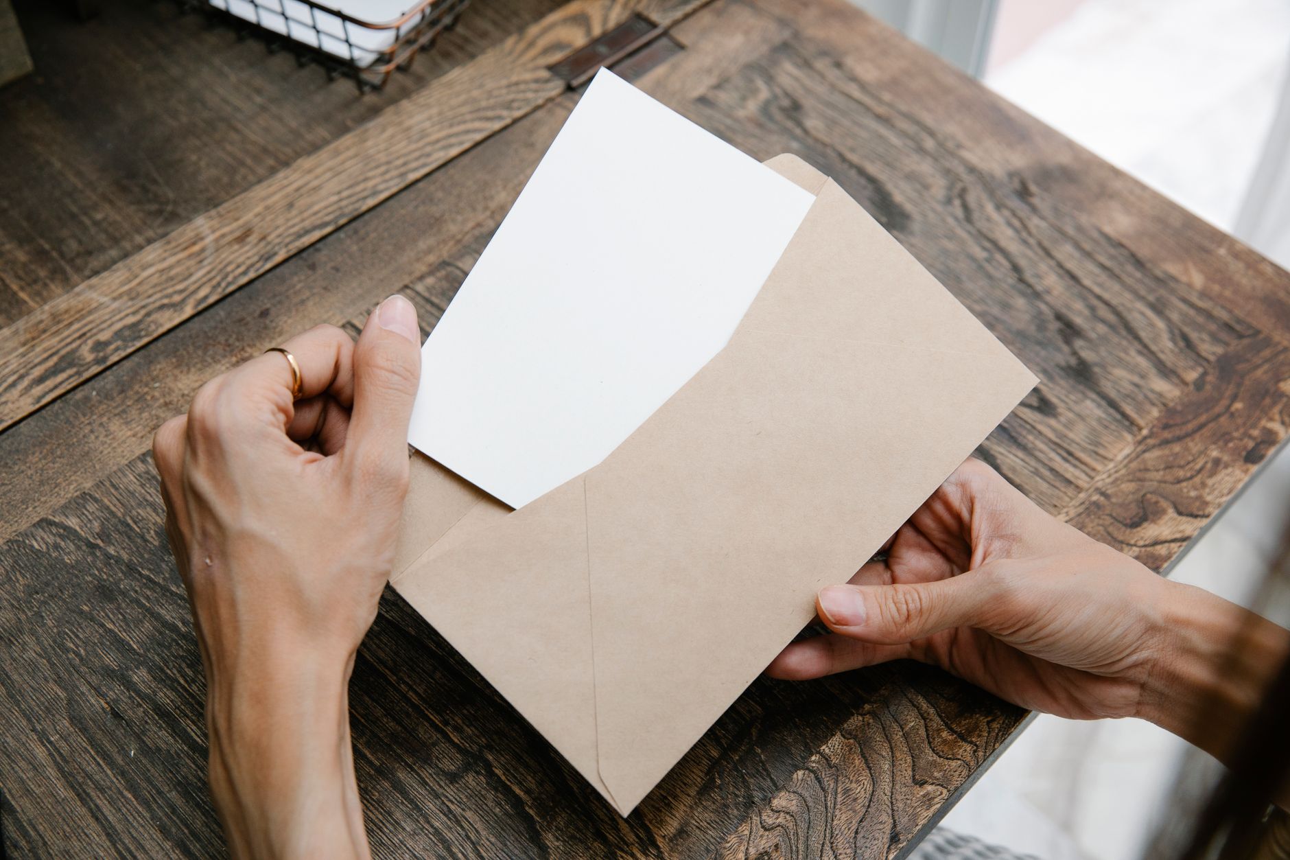 Two hands lift a folded letter out of a brown envelope on a wooden table.