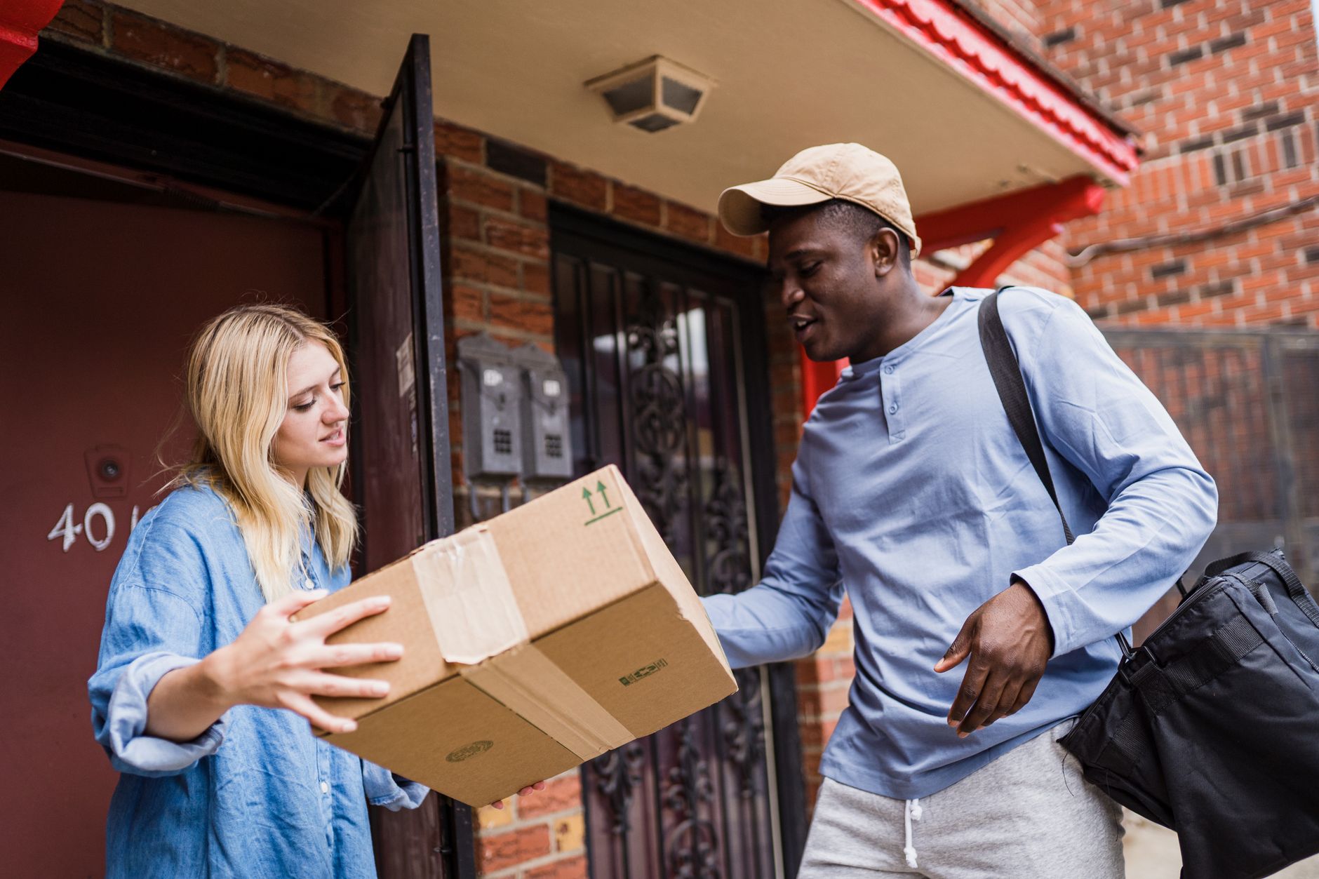 A delivery driver hands a cardboard parcel to a woman at the front door of a brick house.