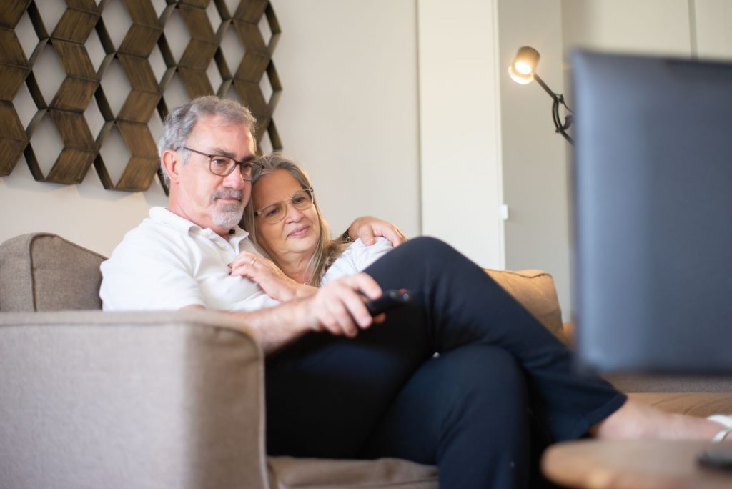 An older couple sit together on a sofa watching television, one of them holding the remote.