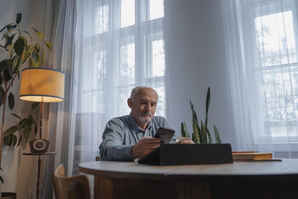 An older man sits at a table by a window at home, holding a phone and looking at a tablet.