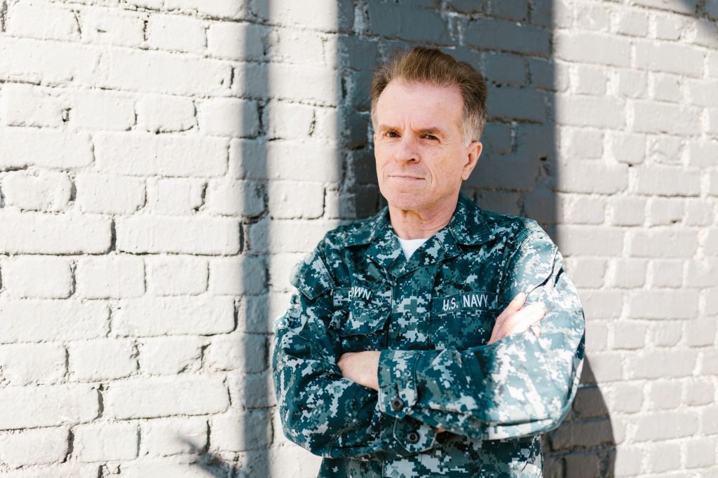 A senior US Navy veteran in uniform stands with arms folded against a painted brick wall.