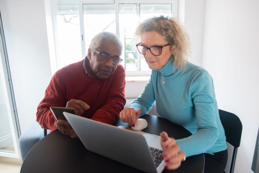 An older couple sit together at a table looking closely at a laptop screen.